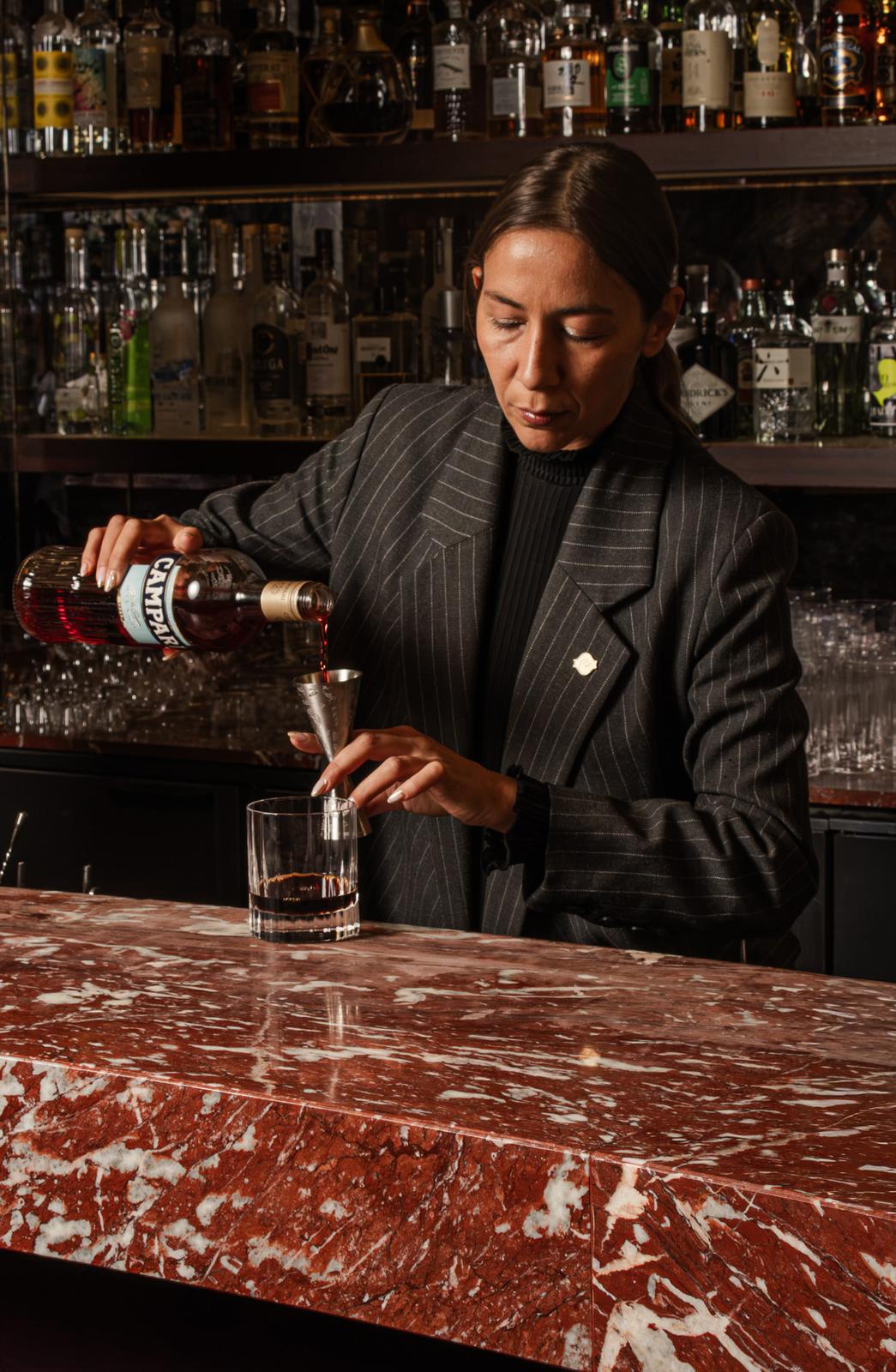 Dosage du Campari au jigger au-dessus d'un verre à whisky, devant l'étagère de spiritueux du bar Joy