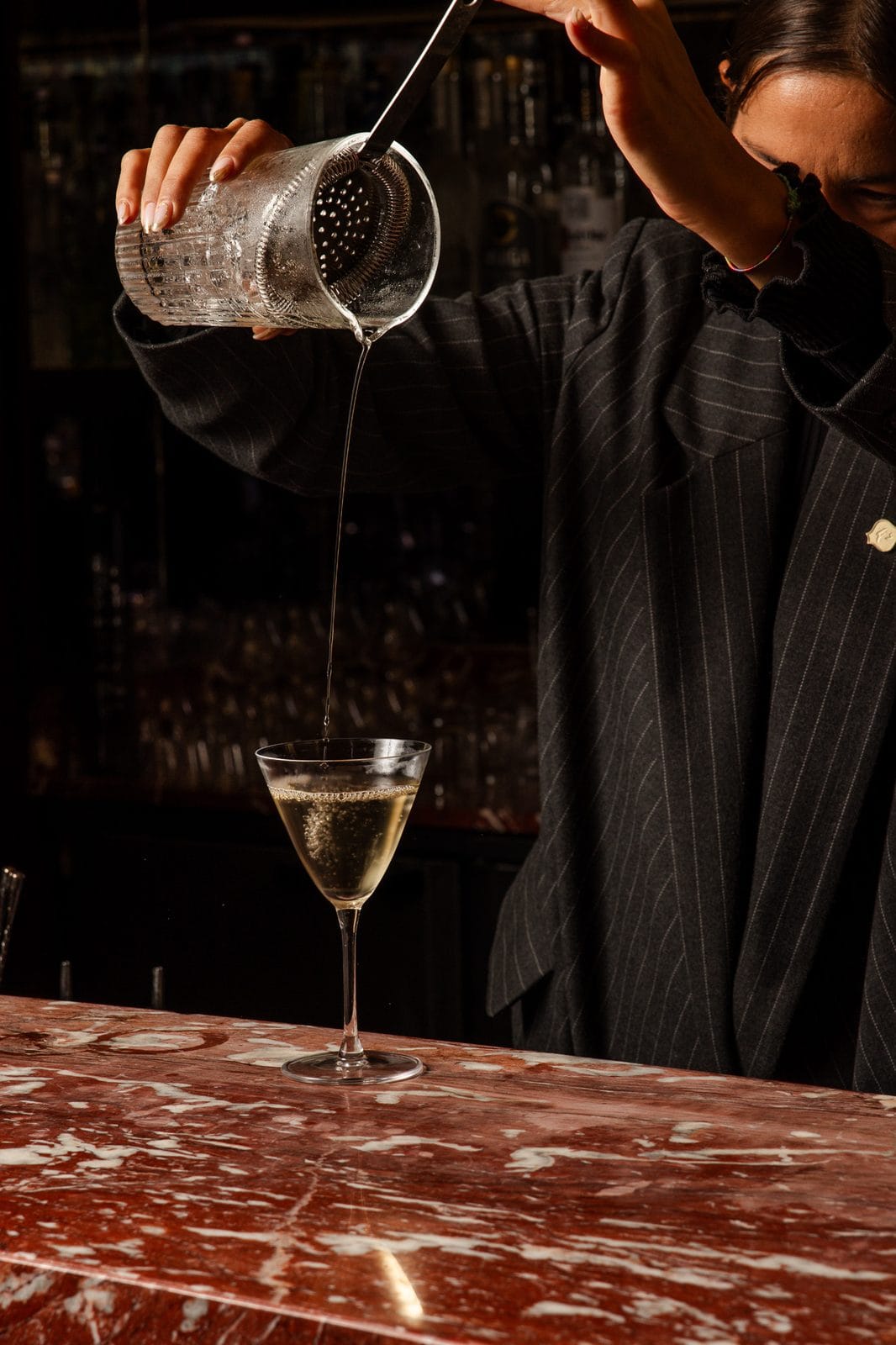 Cocktail clair filtré depuis un mixing glass dans un verre à pied conique, au comptoir de marbre rouge