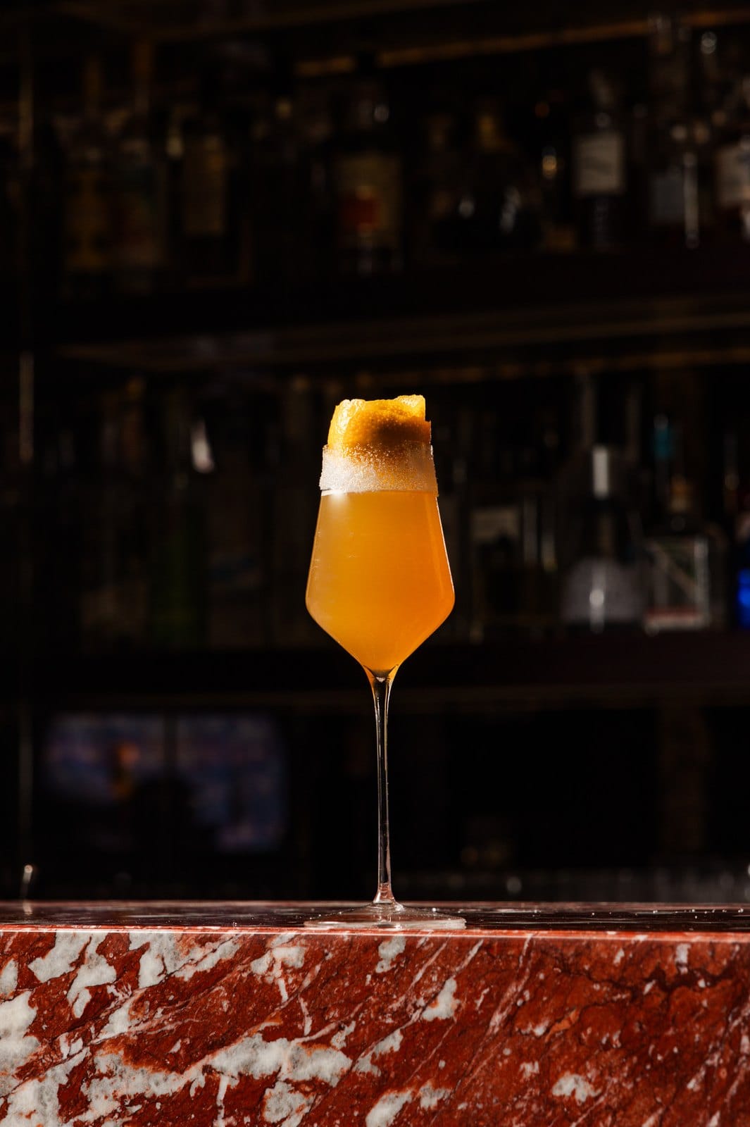 Cocktail orangé en verre à pied surmonté d'un zeste, sur le bord du comptoir de marbre rouge