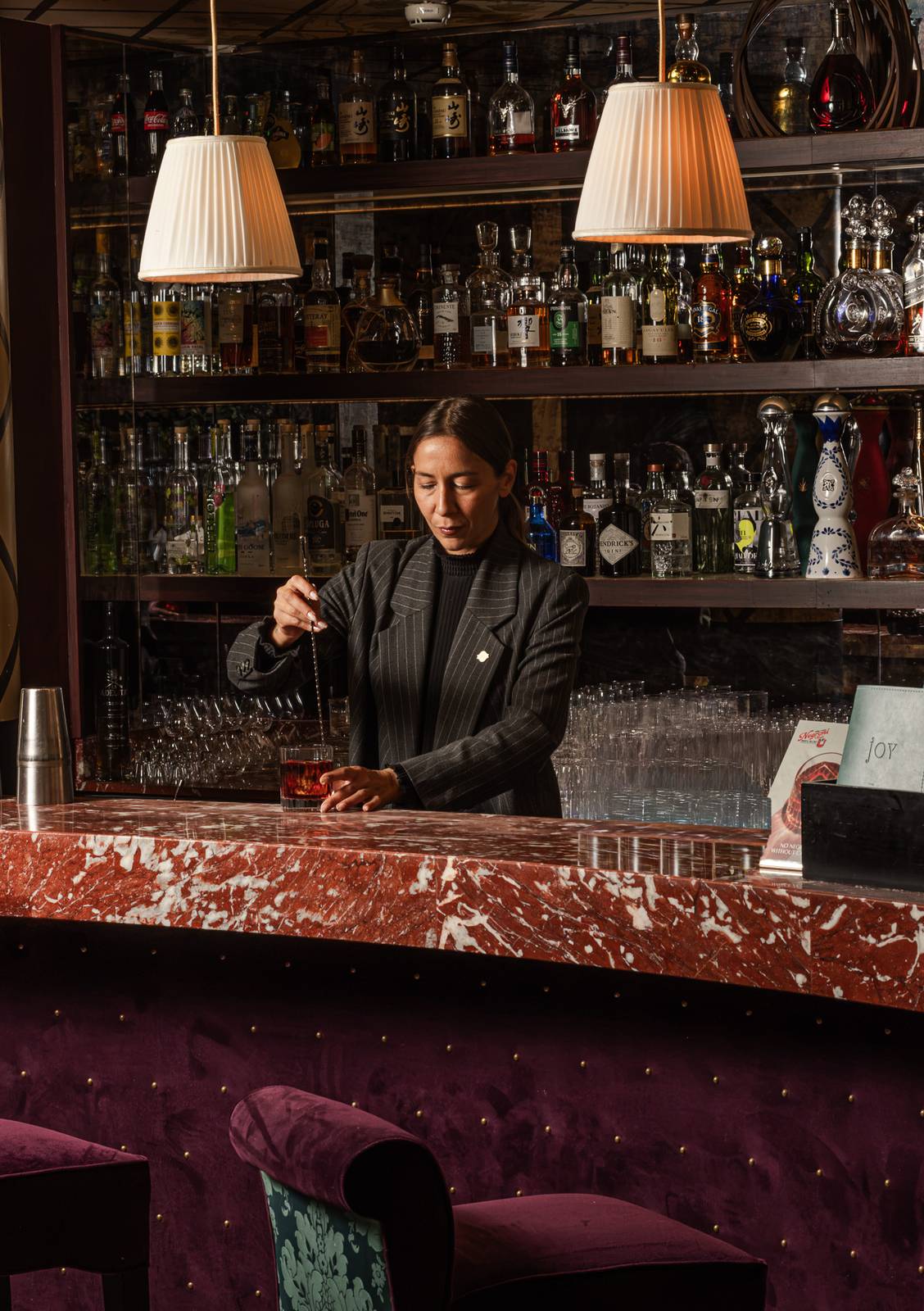 Bartender remuant un Negroni au comptoir de marbre rouge du bar Joy, sous deux abat-jour plissés, devant l'étagère de bouteilles, Fouquet's Paris