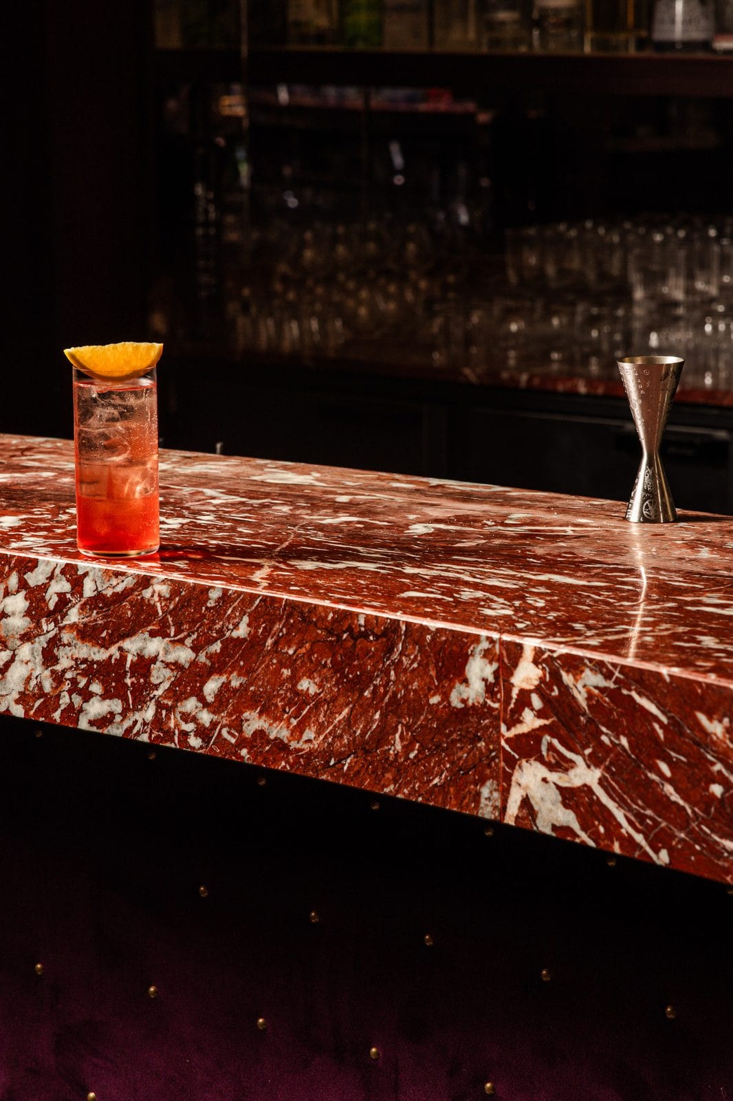 Long drink rouge sur glace avec demi-tranche d'orange, et jigger gravé au bout du comptoir de marbre