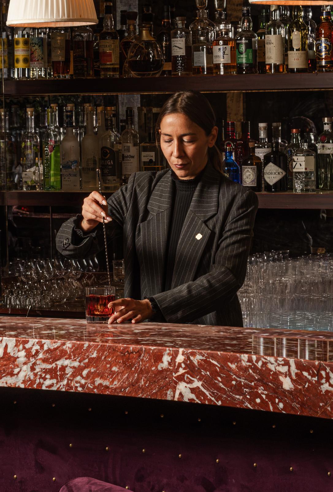 Bartender en tailleur à fines rayures remuant un Negroni à la cuillère de bar, comptoir de marbre rouge et velours prune