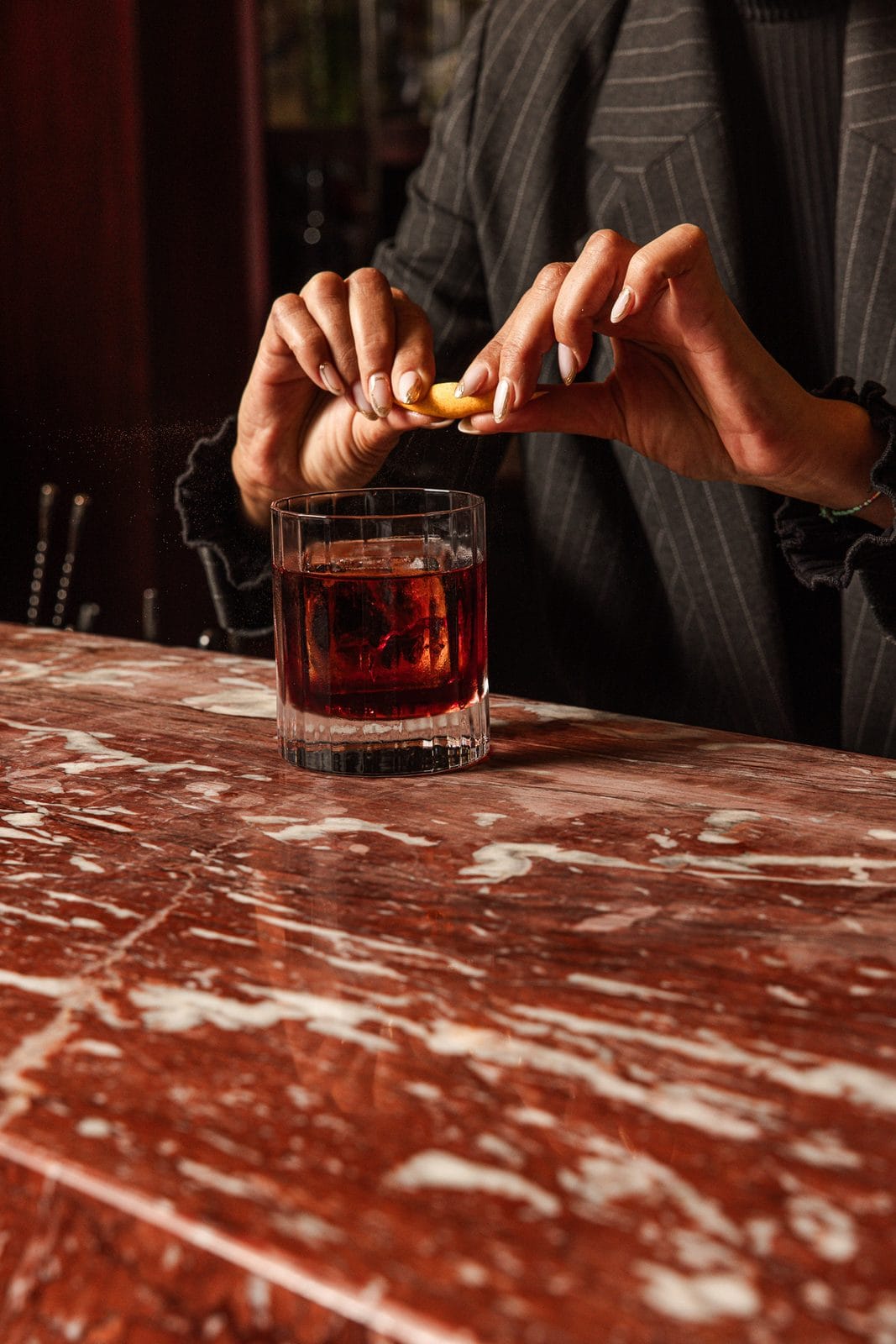 Negroni sur gros glaçon en verre cannelé, zeste d'orange exprimé au-dessus du comptoir de marbre rouge