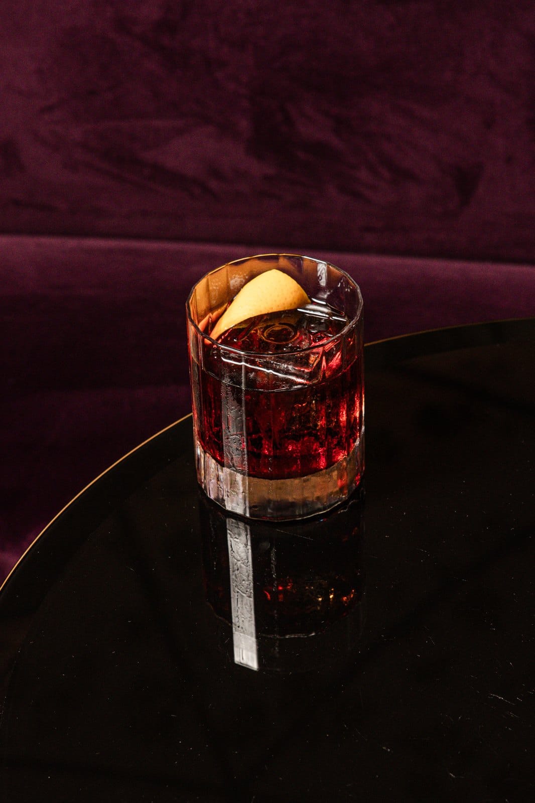 Negroni au zeste d'orange sur un guéridon laqué noir, devant une banquette de velours prune