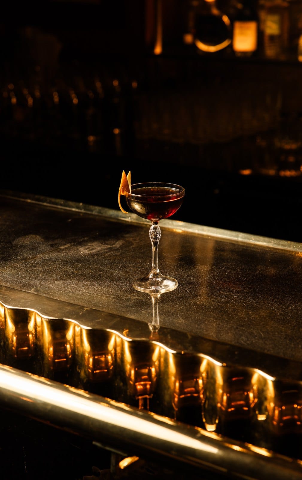 Cocktail ambré en coupe ciselée, double zeste d'orange, sur un comptoir de métal dans une lumière dorée